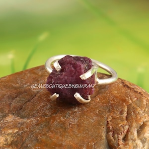 May include: A silver ring with a raw, pink ruby gemstone. The ruby is set in a claw setting and is surrounded by a silver band.