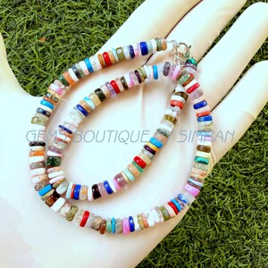 May include: A multi-colored beaded necklace featuring small, flat, circular gemstone discs in various hues including red, blue, white, green, and pink. The necklace is secured with a silver-toned metal lobster clasp and features the text GEMS BOUTIQUE SIMRAN.