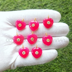 Pode incluir: Uma coleção de oito pingentes em forma de coração rosa brilhante com laços dourados. Cada coração apresenta uma gema redonda transparente central. Os pingentes são exibidos em uma superfície branca sobre um fundo verde.