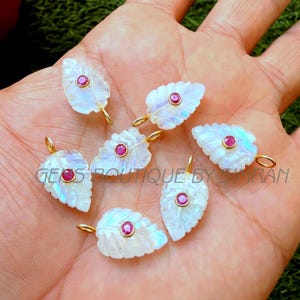 May include: Seven leaf-shaped pendants with a carved design. Each pendant is white with iridescent hues, a small red gemstone, and a gold-colored loop. The pendants are displayed on a hand.