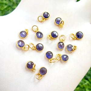May include: A collection of small, round, purple gemstone charms with gold-colored metal settings and loops. The charms are scattered across a surface, showcasing their polished appearance and potential for jewelry making.