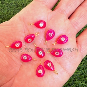 May include: A collection of hot pink teardrop-shaped pendants with a gold-coloured loop and a clear, round gem in the centre. The pendants are displayed on a hand, with the text "GEMS BOUTIQUE BY IMRAN" visible.