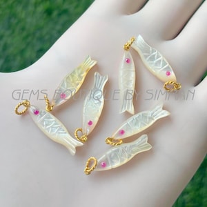 Può includere: Sette ciondoli iridescenti a forma di pesce con anelli color oro e accenti occhi rosa. I ciondoli hanno un motivo a scaglie intagliate e sono disposti su uno sfondo chiaro. Il testo "GEMS BOUTIQUE BY SIMAN" è visibile.