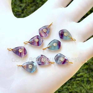 Può includere: Una collezione di ciondoli a forma di goccia con un anello color oro nella parte superiore. Ogni ciondolo presenta una pietra sfaccettata multicolore con una gemma trasparente rotonda al centro. Le pietre mostrano sfumature di viola, blu e trasparente.