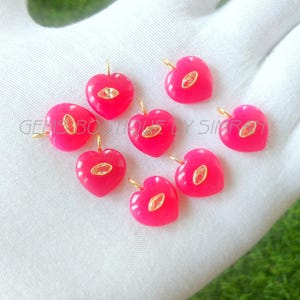 Może przedstawiać: Kolekcja jasnoróżowych zawieszek w kształcie serca ze złotymi krawędziami i centralnym owalnym akcentem. Zawieszki ułożone są na białej powierzchni. Widoczny jest tekst "GEMS BOUTIQUE BY SIMROY".