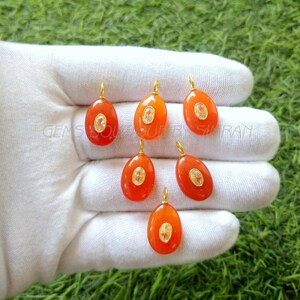 Puede incluir: Siete colgantes ovalados de piedra naranja con detalles dorados y detalles de cristal transparente. Los colgantes están sobre una superficie blanca.