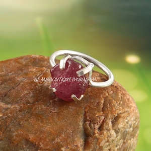 Könnte beinhalten: Ein silberner Ring mit einer Krappenfassung, in der ein roher, rosafarbener Rubin-Edelstein sitzt. Der Ring liegt auf einem braunen Stein.