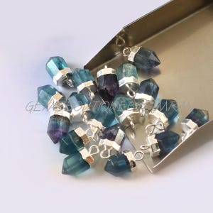 May include: A group of 18 silver-toned gemstone pendants with a faceted, pointed shape. The gemstones are a mix of blue and green colors with a slight purple hue. Each pendant has a silver loop for attaching to a chain or cord.