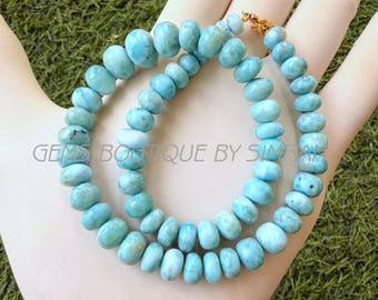 May include: A necklace composed of round, light blue Larimar beads with visible white marbling and dark inclusions. The beads are strung together and secured with a gold-toned metal clasp. The text GEMS BOUTIQUE BY SIMRAN is visible on the image.