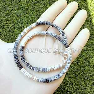 May include: A beaded necklace featuring small, flat, disc-shaped beads in varying shades of white, gray, and black with a mottled pattern. The necklace includes a silver-toned metal lobster claw clasp and an extender chain. Text reads GEMS BOUTIQUE BY SIMRAN.