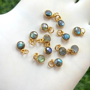 Puede incluir: Una colección de pequeños colgantes redondos de piedra labradorita con aros dorados. Las piedras preciosas muestran tonos iridiscentes azules, verdes y grises. Los colgantes están dispuestos sobre una superficie blanca, lo que sugiere suministros para la fabricación de joyas.