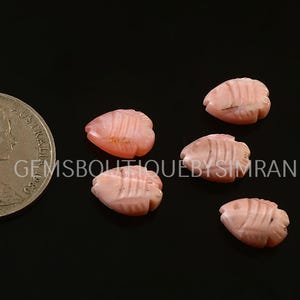Peut inclure: Cinq perles roses sculptées en forme de poisson sont présentées sur fond noir. Les perles présentent des lignes sculptées détaillées. Une pièce de monnaie est dans l'image pour référence de taille.