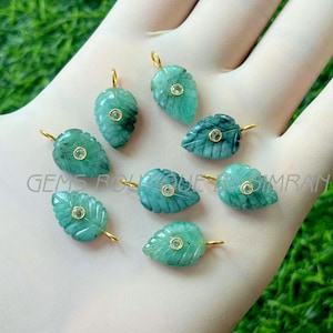 Peut inclure: Plusieurs pendentifs en forme de feuilles vert émeraude avec des bélières dorées. Chaque pendentif est orné d'une petite pierre précieuse ronde et transparente au centre. Les pendentifs sont présentés sur une surface claire.