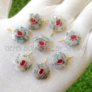 May include: A collection of gemstone pendants. Each pendant features a hexagonal, speckled, gray and white stone with a vibrant red oval gemstone in the center. The pendants have gold-colored loops for hanging. The text "GEMS BOUTIQUE BY SIMRAN" is visible.