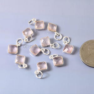 Peut inclure: Plusieurs pendentifs carrés en quartz rose avec des boucles argentées, dispersés sur une surface bleu clair. Les pendentifs sont d'une teinte rose pâle. Une pièce de monnaie est visible en bas à droite.