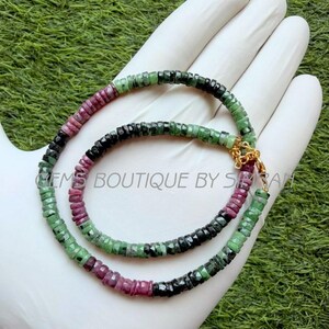 May include: A necklace composed of small, faceted, disc-shaped beads in alternating shades of deep pink, forest green, and black. The strand features a gold-toned metal lobster clasp closure and includes the text GEMS BOUTIQUE BY SIMRAN.