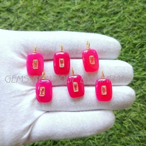 Peut inclure: Sept pendentifs en pierres précieuses fuchsia avec des incrustations rectangulaires en pierre transparente et des bélières dorées. Les pendentifs sont présentés sur un gant blanc sur fond d'herbe verte. Le texte "GEMS BOUTIQUE" est visible.