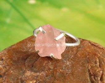 May include: A silver ring featuring a rough-cut, light pink gemstone. The stone is irregularly shaped and set in a simple, modern band. The ring is displayed on a brown surface against a blurred green background. The text "GEMSBOUTIQUEBYBIMA" is visible.