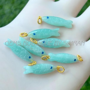 Pode incluir: Uma coleção de pingentes em forma de peixe azul claro com laços dourados e olhos azuis. Os pingentes são esculpidos com detalhes e parecem ser feitos de um material translúcido. O texto "GEMS BOUTIQUE BY SIMRAN" é visível.