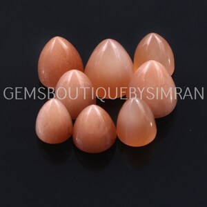 Puede incluir: Un grupo de ocho piedras preciosas cabujón en forma de lágrima de color melocotón con una parte superior puntiaguda. Las piedras preciosas están dispuestas en círculo sobre una superficie negra.