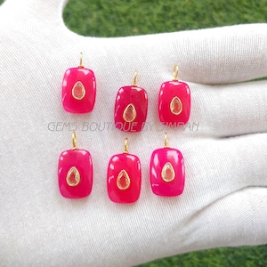 May include: Six rectangular, fuchsia-coloured pendants with gold-coloured bails. Each pendant features a central, oval-shaped, gold-coloured stone. The pendants are displayed on a white surface. The text "GEMS BOUTIQUE BY SIMPAN" is visible.