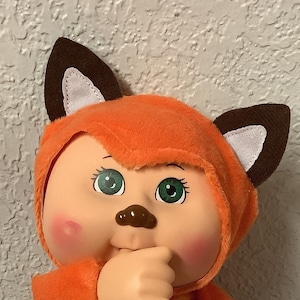 May include: A plush toy fox with orange fur, brown ears, and a yellow and orange striped tummy. The toy has a round face with green eyes and a brown nose.