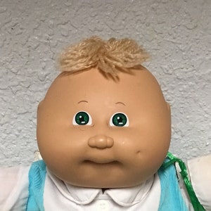 Può includere: Una bambola Cabbage Patch Kid con capelli biondi, occhi verdi e un incarnato rosato. La bambola indossa un vestito blu e bianco con un elefante ricamato sul davanti.