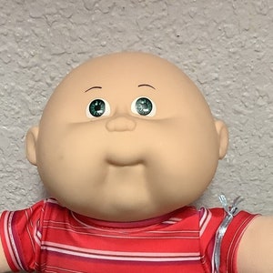 May include: A bald Cabbage Patch Kid doll wearing a red and white striped shirt, blue jeans, and white socks. The doll is holding a light blue mesh bag.
