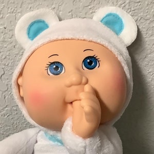 Pode incluir: Uma boneca de pelúcia Cabbage Patch Kid branca vestindo uma roupa de inverno azul e branca com um padrão de flocos de neve azuis e brancos. A boneca está usando um chapéu branco com orelhas de urso azuis e está colocando o polegar na boca. A boneca tem olhos azuis e bochechas rosadas. A boneca está sentada em um cobertor colorido com uma etiqueta CPK presa ao seu pé.