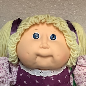 Puede incluir: Muñeca vintage con cabello rubio de lana en coletas, ojos azules y rostro redondo. La muñeca viste un vestido floral morado y blanco con lazo a juego, calcetines de encaje blanco y zapatos blancos. Se ve una bolsa verde.