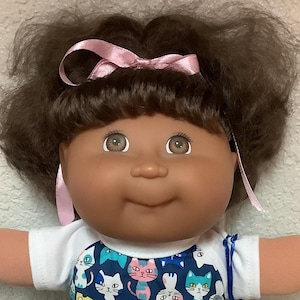 May include: A vintage doll with brown hair styled in pigtails, a pink bow, and brown eyes. The doll wears a white shirt and a blue dress with a cat pattern. White shoes and a blue bag complete the outfit.