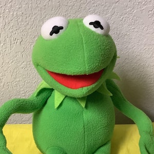 以下が含まれることがあります： 緑色のぬいぐるみ、カーミット・ザ・フロッグのパペット。大きく笑っていて、白い目が大きく開いています。パペットは黄色の表面に座っています。