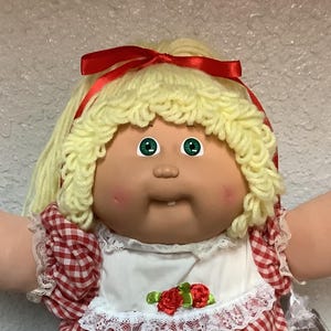May include: A vintage doll with blonde yarn hair and green eyes, wearing a red and white checkered dress with a white lace collar. A red bow adorns the doll's hair. The doll is wearing white shoes with lace trim.