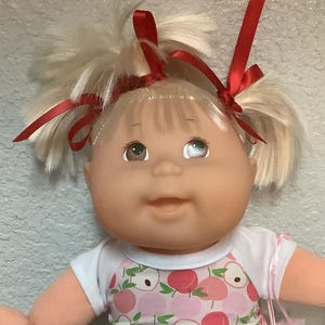 May include: A vintage doll with blonde pigtails tied with red ribbons. The doll wears a white and pink peach-patterned romper, pink socks, and carries a pink pacifier in a small bag. The doll has a smiling expression.