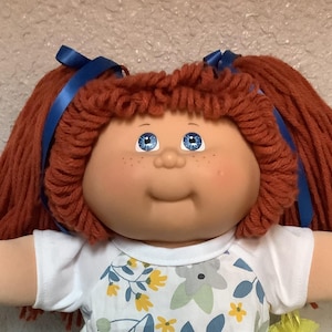 May include: A vintage doll with red yarn pigtails tied with blue ribbons. The doll has blue eyes, freckles, and a smiling expression. It wears a white dress with a floral pattern and white shoes with lace trim. A yellow tag is attached.