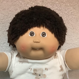 May include: A Cabbage Patch Kid doll with brown hair, rosy cheeks, and a small smile. The doll wears a white shirt with koala bear prints, green trousers, and white trainers with blue stripes. A small bag with an orange object hangs from its arm.