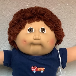 Könnte beinhalten: Eine Vintage-Puppe mit rundem Gesicht, braunem Lockenhaar und rosigen Wangen. Sie trägt ein marineblaues T-Shirt mit einem roten Feuerwehrauto-Motiv, eine rote Hose und weiße Turnschuhe mit blauen Akzenten. Die Arme der Puppe sind ausgestreckt.