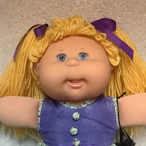 May include: A vintage Cabbage Patch Kid doll with blonde yarn hair styled in pigtails, adorned with purple ribbons. The doll wears a purple dress with floral print, pockets, and white lace socks.