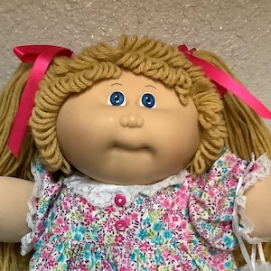 May include: A vintage doll with blonde yarn hair styled in pigtails, adorned with pink ribbons. The doll wears a floral print dress with a white lace collar and cuffs, and white shoes with lace trim. The doll has blue eyes and a neutral expression.