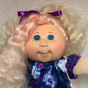 Peut inclure: Une poupée Cabbage Patch Kids vintage avec des cheveux blonds bouclés, des reflets roses et des yeux bleus. La poupée porte une robe violette et bleue imprimée galaxie, des chaussettes en dentelle blanche et des chaussures violettes. Un nœud violet orne les cheveux de la poupée.