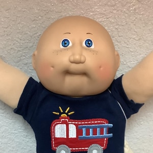 May include: A Cabbage Patch Kid doll wearing a navy blue shirt with a red and white fire truck design, red pants, and white shoes. The doll has a bald head, blue eyes, and rosy cheeks. The doll's arms are outstretched.