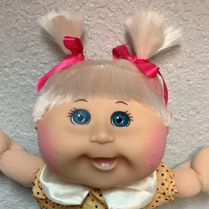 Peut inclure: Une poupée vintage avec des couettes blondes ornées de rubans roses. La poupée a les yeux bleus, les joues roses et porte un bloomer jaune à pois avec un col blanc et un nœud jaune. La poupée est debout, les bras levés.