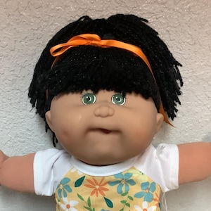 May include: A vintage doll with black hair, green eyes, and an orange ribbon. The doll wears a white shirt and a yellow dress with blue and white floral patterns. White shoes and socks complete the outfit. The doll's arms are outstretched.