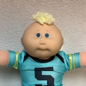 Könnte beinhalten: Cabbage Patch Kids Puppe mit einem türkisfarbenen Football-Trikot mit der Nummer 5, dunkelblauen Shorts und weißen Turnschuhen. Die Puppe hat blondes Haar und blaue Augen. Die Puppe steht mit ausgestreckten Armen da.