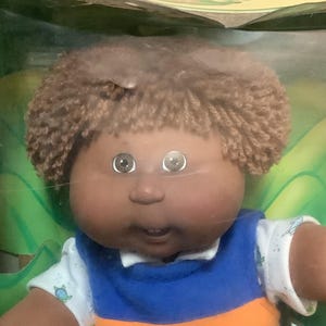May include: Cabbage Patch Kids Percy Larry doll in original packaging. The doll has brown skin, brown curly hair, and wears a blue and orange striped vest, a white shirt, and corduroy trousers. The packaging features the Cabbage Patch Kids logo.