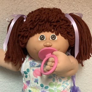 May include: A vintage doll with brown yarn hair styled in pigtails with lavender ribbons. The doll wears a floral print dress, purple shorts, white lace socks, and white shoes. It holds a pink pacifier.