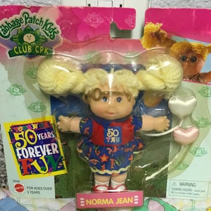 May include: A Cabbage Patch Kids Club CPK doll called Norma Jean. The doll is wearing a blue and red dress with a white collar and a white headband. The doll is holding a pink heart-shaped balloon. The doll is packaged in a pink and green box with the words "50 Years Forever Fun" on it.