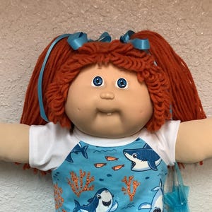 Puede incluir: Muñeca Cabbage Patch Kid con cabello rojo de hilo en coletas, ojos azules y una expresión sonriente. La muñeca viste una camisa azul con gráficos de tiburones, pantalones cortos blancos con encaje y zapatos blancos. Un accesorio azul se sostiene en su mano.