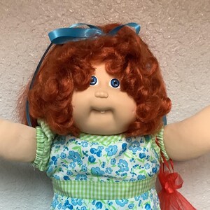 May include: A vintage doll with red curly hair and blue eyes, wearing a blue floral dress with a green gingham waist and white lace-trimmed shoes. A blue ribbon adorns the doll's hair, and a red mesh bag hangs from its arm.