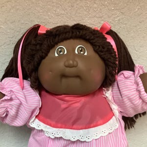 Peut inclure: Une poupée Cabbage Patch Kids vintage avec des couettes brunes ornées de rubans roses. La poupée porte une robe rayée rose et blanche avec une bordure en dentelle et des chaussures roses assorties. La poupée a un teint foncé et des yeux bruns.